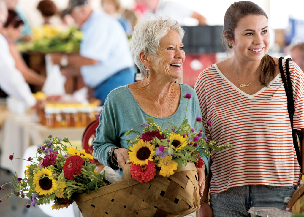 Two women with flowers smile at a farmers market.